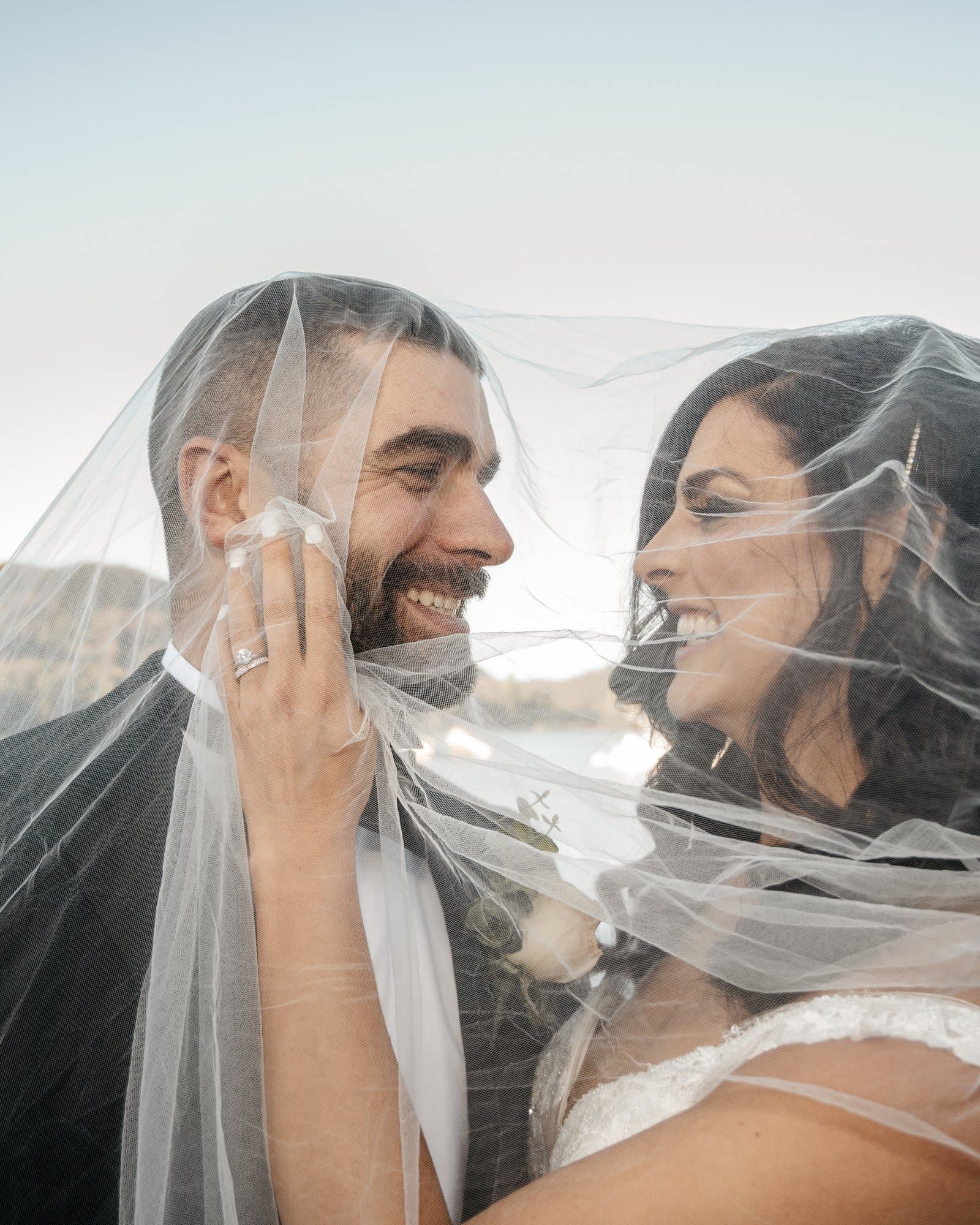 Bride and groom laughing together under her veil at the edge of a lake