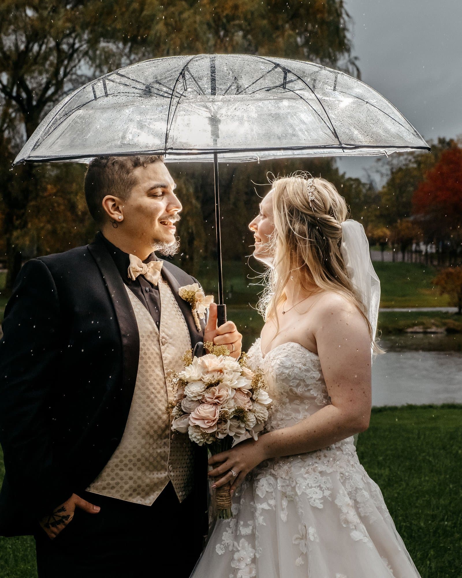 Bride and groom laughing under a clear umbrella in the rain by a pond