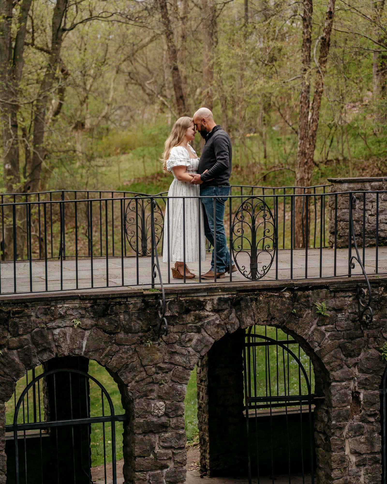 Engaged couple forehead to forehead on a stone arch bridge with wrought-iron railings