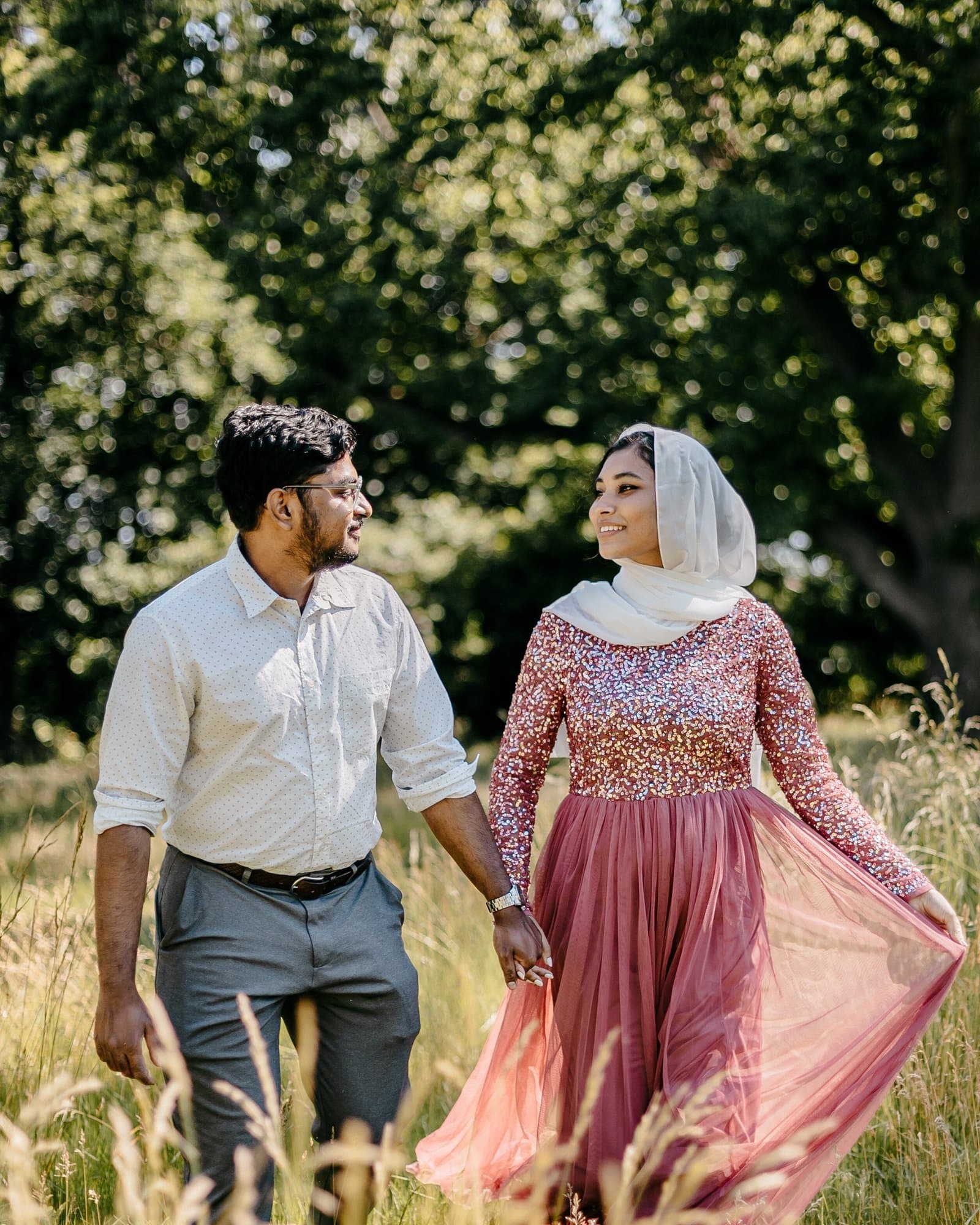 Couple holding hands walking through a golden meadow, her sequined rose dress catching the light