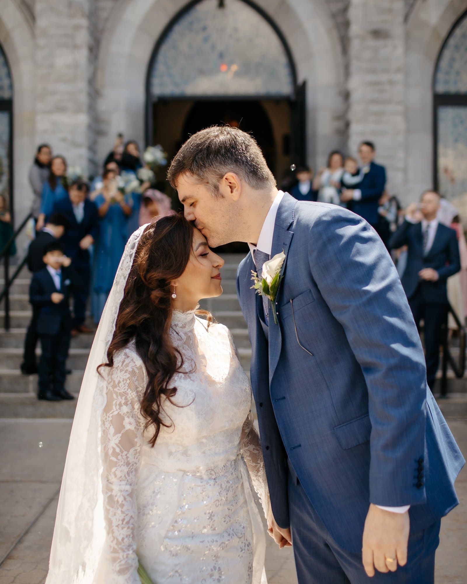 Groom kissing the bride's forehead on stone church steps as guests celebrate