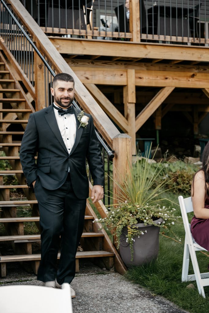 A groom wears a black tuxedo and a white flower and walks down the aisle after paying the average cost of a wedding in ny